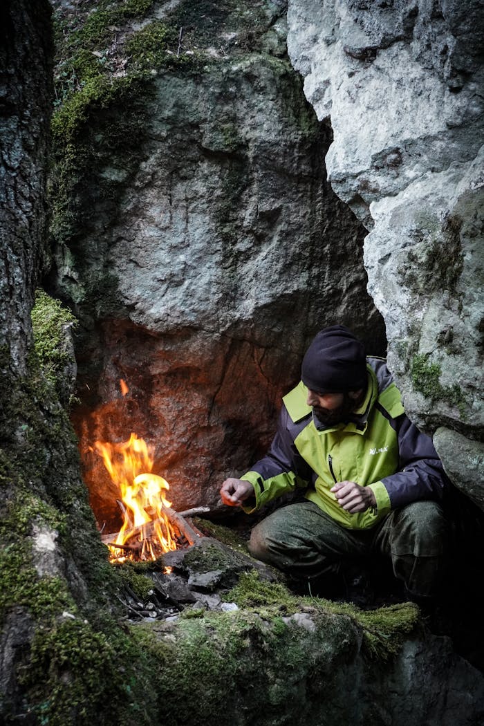 Home Man in green jacket lights campfire in rocky mountain shelter for survival.